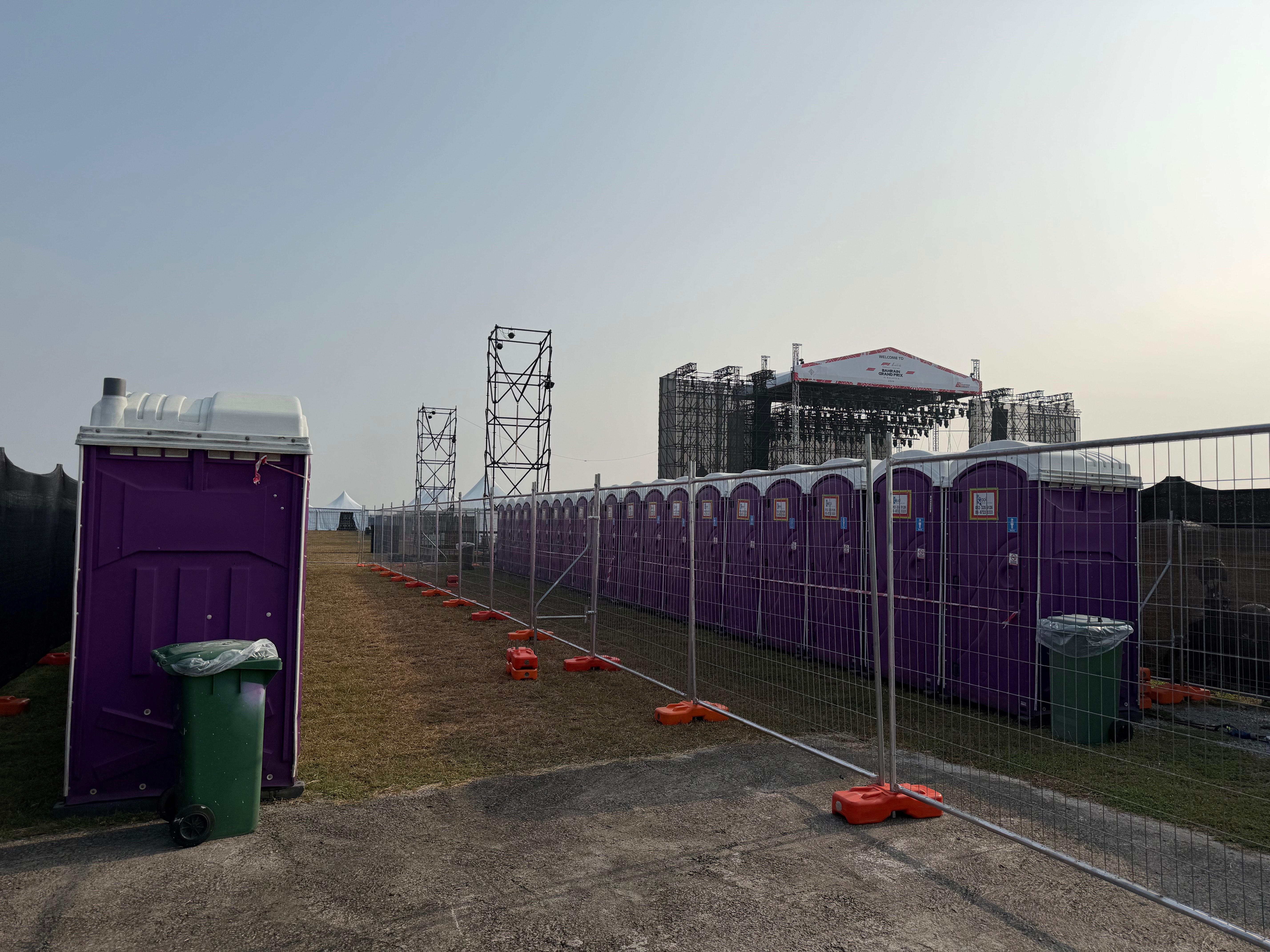 Long fenced row of purple EQool portable toilets with waste bins and the Bahrain Grand Prix stage behind