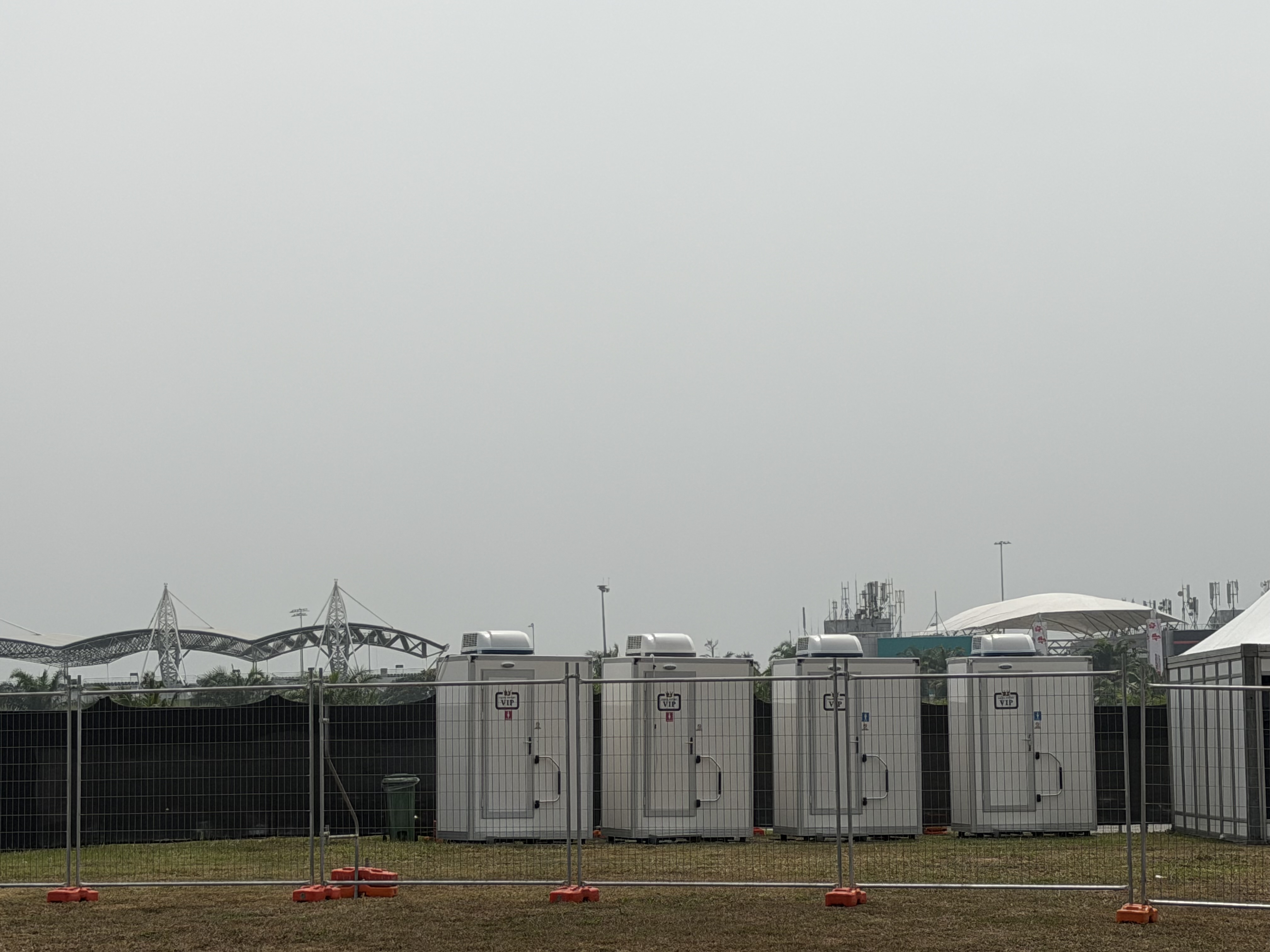 Row of four white EQool VIP cabin toilets behind temporary fencing with the circuit grandstand roof behind
