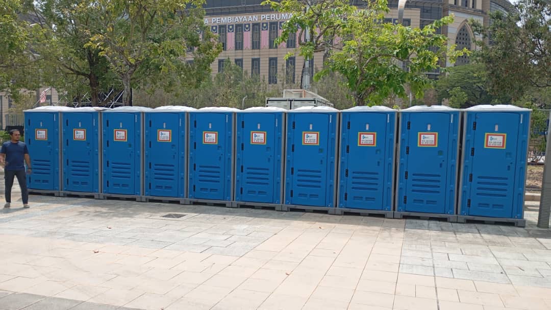 Ten blue EQool portable toilets in a row on a paved plaza with a crew member standing beside them