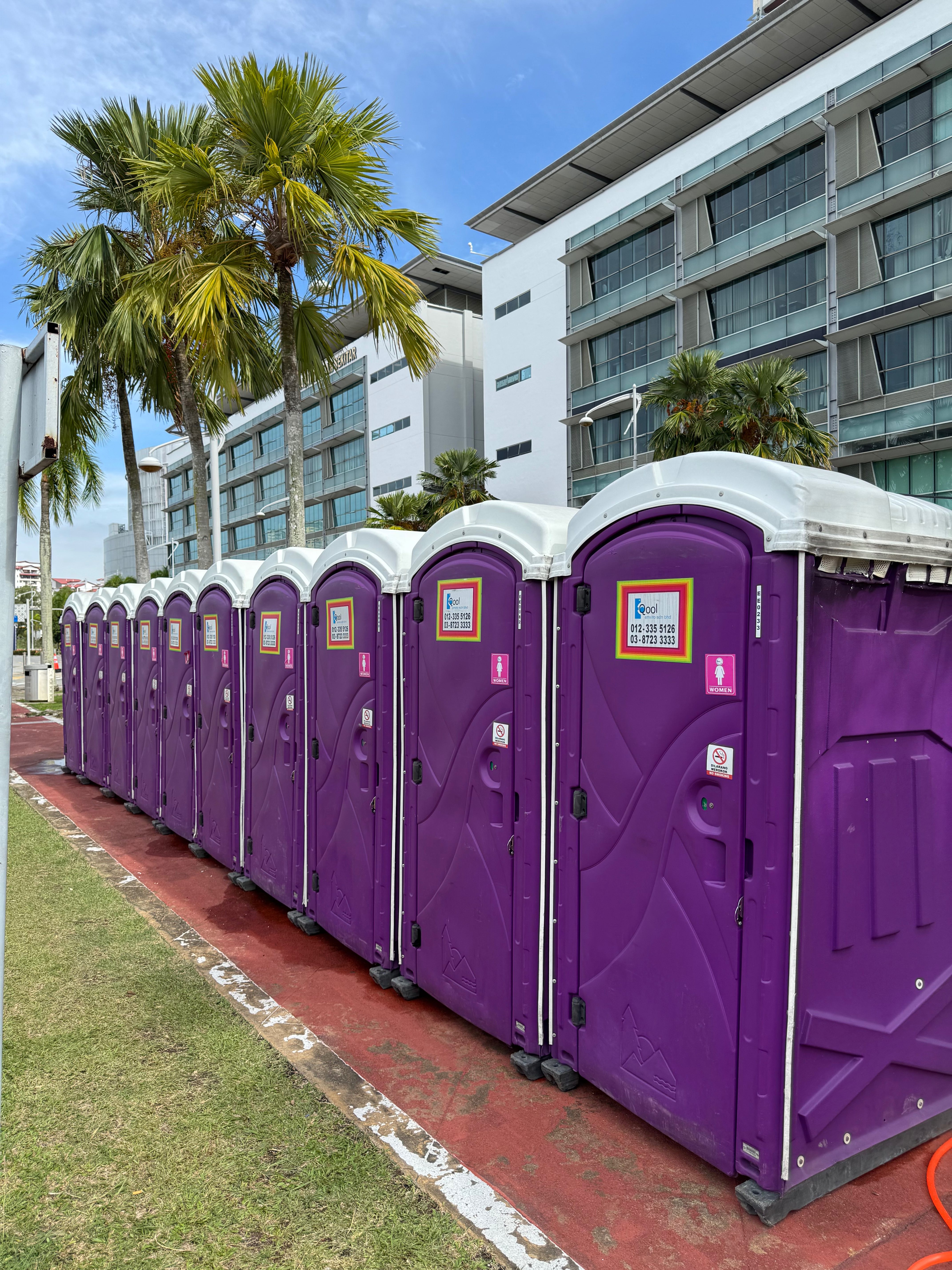 Purple EQool portable toilets marked with women signs, lined up along a red track with palm trees behind