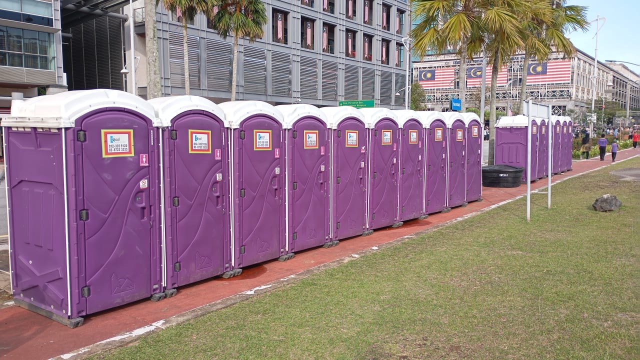 Purple EQool portable toilets set up along a grass verge with Malaysian flags on the buildings behind
