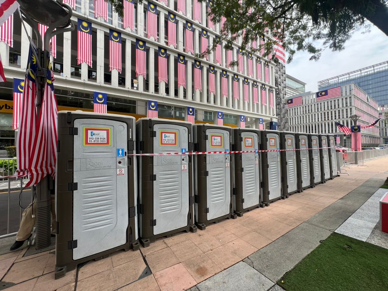 Row of grey and brown EQool portable toilets with a safety tape line under rows of Jalur Gemilang banners
